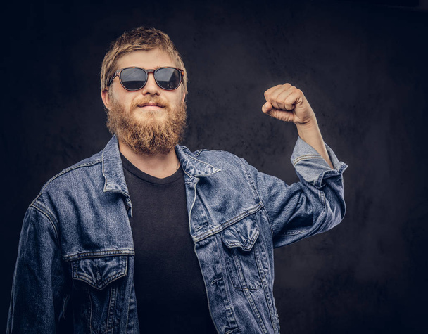 Pensive hipster guy wearing sunglasses dressed in jeans jacket shows how strong he is on a dark background. - Photo, Image