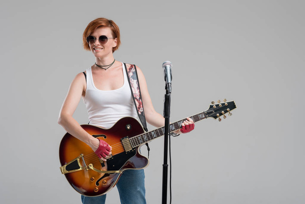 Portrait of a beautiful young rock woman in sunglasses with a guitar. She sings a song in studio microphone - Foto, Imagem