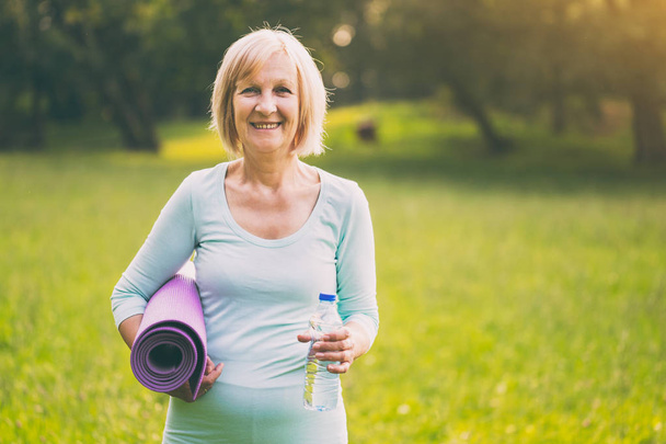 Portrait of sporty senior woman holding bottle of water and mat in the nature.Image is intentionally toned. - Foto, Imagen