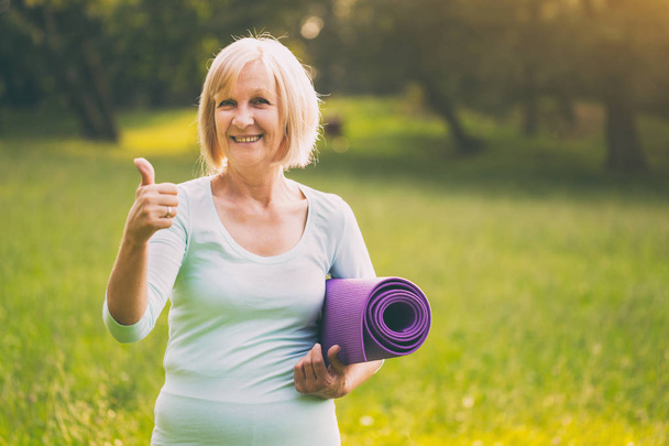Portrait of sporty senior woman showing thumb up and holding mat in the nature.Image is intentionally toned. - Foto, Imagen