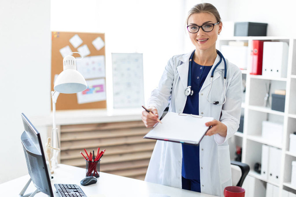 A young girl in a white coat is standing near a table in her office and holding a tablet and a pen. A stethoscope hangs around her neck. - Foto, Imagem