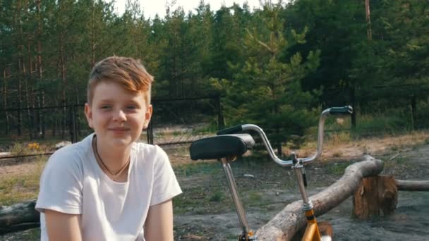 Χαριτωμένο ξανθιά με γαλανά μάτια ένα έφηβο αγόρι κάθεται και χαμόγελο σε ένα πάρκο δίπλα στο ποδήλατο και συνομιλίες για το καλοκαίρι. Συναισθηματικό πορτραίτο ενός αγοριού - Πλάνα, βίντεο