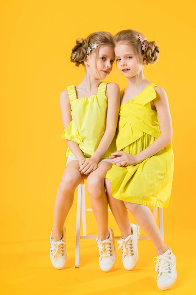 Δίδυμα κορίτσια κάθονται σε μια καρέκλα σε κίτρινο φόντο. - Φωτογραφία, εικόνα
