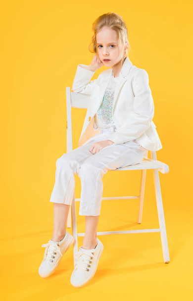 A teenage girl in white clothes is sitting on a chair on a yellow background. - Fotografie, Obrázek