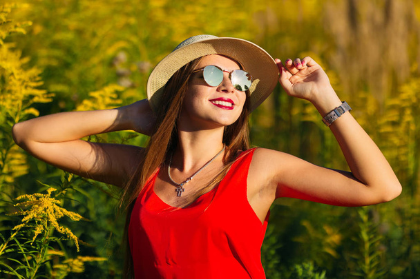 девушка в стильной шляпе и солнцезащитных очках мило улыбается и стоит среди высокой травы
 - Фото, изображение