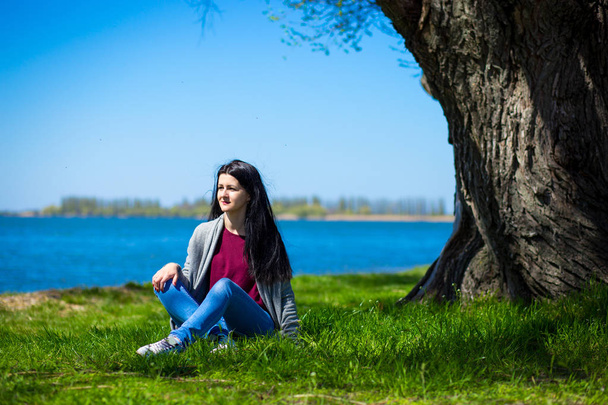 Mulher jovem bonita em jeans senta-se na margem do rio perto de árvore grande em dia ensolarado - Foto, Imagem