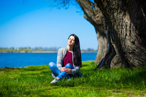 Hermosa mujer joven en jeans se sienta en la orilla del río cerca de un gran árbol en un día soleado - Foto, imagen