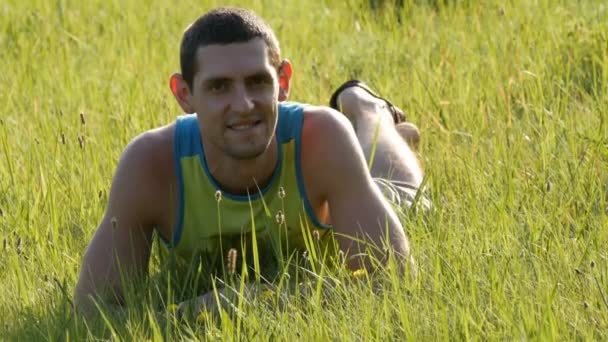 Vrolijke jongeman zit op een groen gras in de zomer en glimlacht - Video