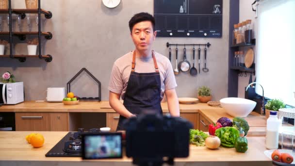 Asijský mladík v kuchyni nahrávání video na kameře. S úsměvem Asiat pracuje na jídlo blogger koncepci s ovocem a zeleninou v kuchyni. - Záběry, video