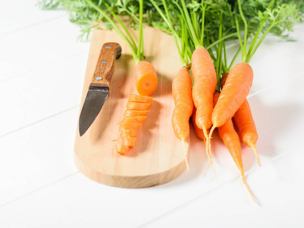 Möhrenscheiben mit einem Messer auf einem Schneidebrett und ein Bund Möhren auf einem weißen Tisch. das Konzept der vegetarischen Ernährung mit natürlichen Rohstoffen. - Foto, Bild