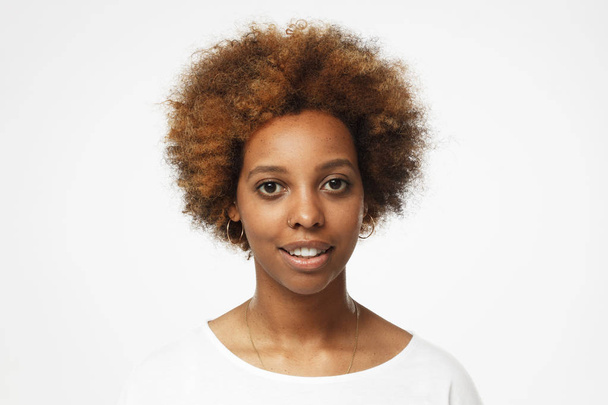 Horizontal headshot of African American female pictured against light grey background with loose curly short hair wearing white T-shirt looking straight at camera with happy smile and interest - 写真・画像