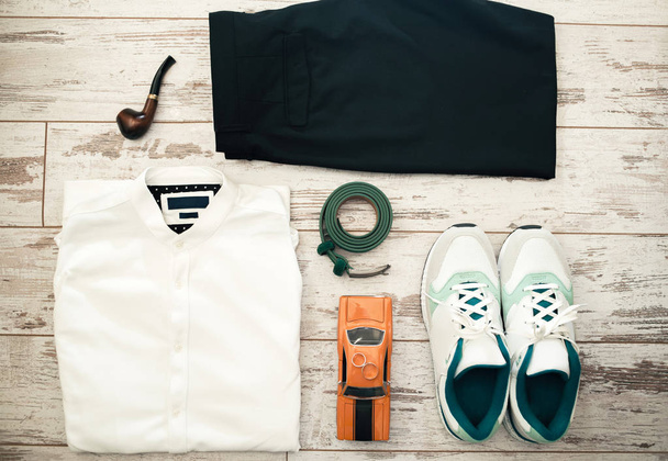 Top view of new man trendy casual clothing on wooden background, white cotton shirt, black pants, belt, sneakers, smoking pipe, toy car and wedding rings. Set of men's casual outfits clothes, trendy fashion, top view - 写真・画像