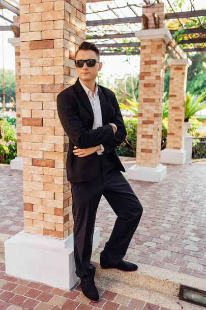 Young stylish handsome man posing in modern city park, wearing suit and sunglasses. - Fotoğraf, Görsel