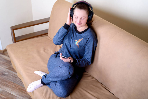 Beautiful young woman sitting on the couch on headphones and listening to music from smart phone. Relaxing on the couch at home - Foto, immagini