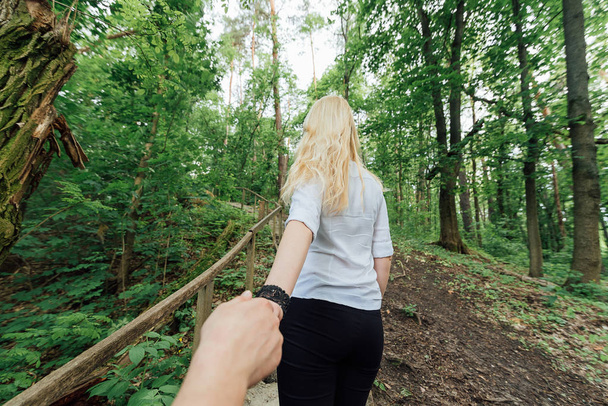 Back view of beautiful young blonde woman in white shirt holding boyfriend's hand outdoors. Girl holding man by hand and leading him to the forest. Follow me. Travel concept - Foto, afbeelding