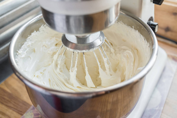 Closeup of electric mixer with whipped smooth dough for cake. Batter being whipped. Mixing white dough in bowl with motor mixer, baking cake - Foto, Imagem
