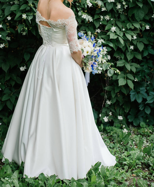 Beautiful bride in wedding dress with wedding bouquet of peonies, blue flowers and greenery near jasmine bush outdoors - Foto, afbeelding