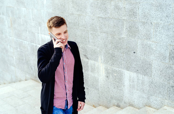 Stylish young man in plaid shirt and jeans walking up stairs and talking on cell phone, full body. Student with smartphone walking up stairs, free space - Photo, image