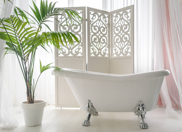 Belle baignoire vide vintage de luxe près d'une grande fenêtre dans l'intérieur de la salle de bain, espace libre. Bain blanc autoportant près de l'écran pliant et du palmier, espace de copie
  - Photo, image