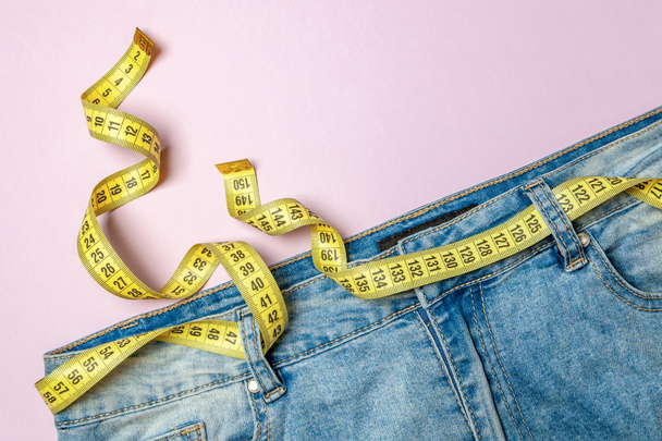 Jeans en gele meetlint in plaats van riem op roze achtergrond. Concept van gewichtsverlies, dieet, detox - Foto, afbeelding