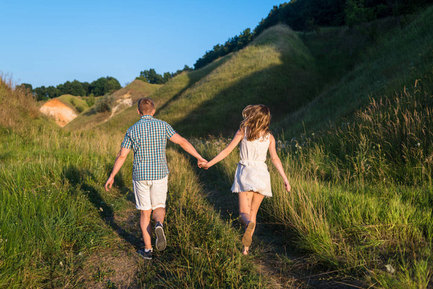 Young loving couple walking on green field near hills, free space. Young beautiful couple of travelers running near hill, back view. Stylish man and woman rest in country side outdoors - 写真・画像