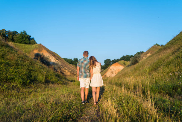 Young loving couple walking on green grass in field between hills, free space. Beautiful couple of travelers enjoy the landscape with blue sky, back view. Stylish man and woman rest in outdoors  - Photo, image