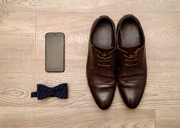 Close up of brown leather shoes men, blue bowtie and mobile phone on wood background, free space. Modern man accessories. Wedding details. Groom accessories. Men casual outfits with shoes and bowtie  - 写真・画像