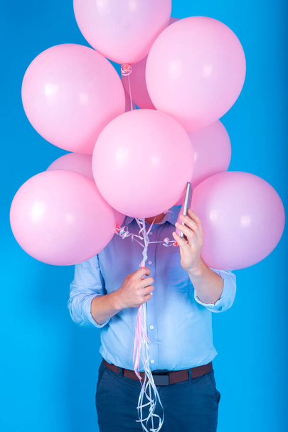 Young handsome man in blue shirt holding pink air balloons and smartphone over blue background, free space for text. Holiday, happy birthday, delivery concept - Φωτογραφία, εικόνα