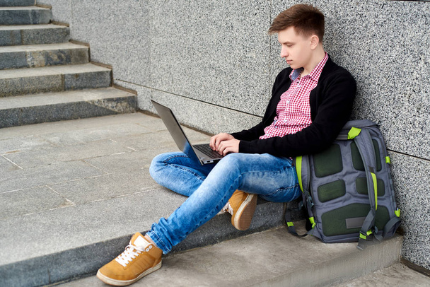 Handsome young man in jacket and jeans with backpack sitting on the stairs under grey stone wall and typing on laptop computer outdoors. Technology and communication concept  - Foto, imagen