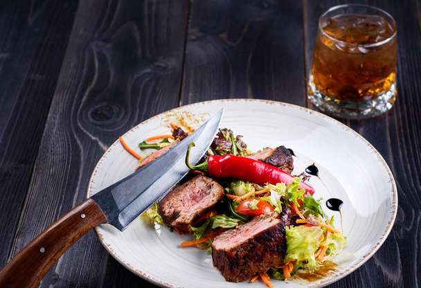 Veal salad with lettuce, carrot, tomato and pepper on white plate on dark wooden background, copy space. Restaurant food, close up. Meat salad and glass of whiskey - Zdjęcie, obraz