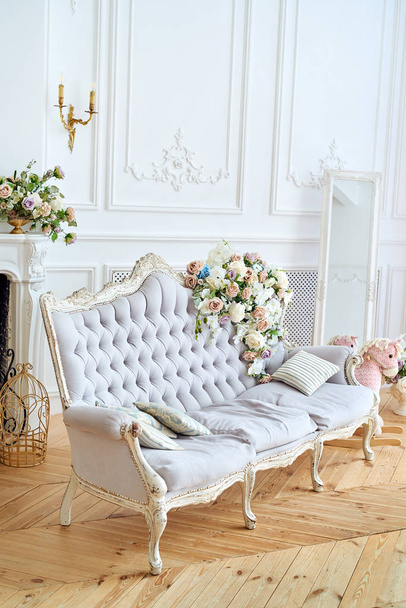 Soft sofa with gray fabric upholstery, pillows and flowers on it, copy space. Luxury rich living room interior design with elegant classic furniture, mirror and fireplace. Vintage textile couch - Фото, изображение