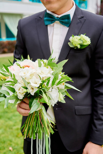 Bruidssuite bruiloft boeket van witte bloemen en groen met linten in bruidegom hand. Bruidegom bruid buiten te wachten. Jonge knappe bruidegom in zwart pak permanent ingedrukt te houden bruiloft boeket - Foto, afbeelding