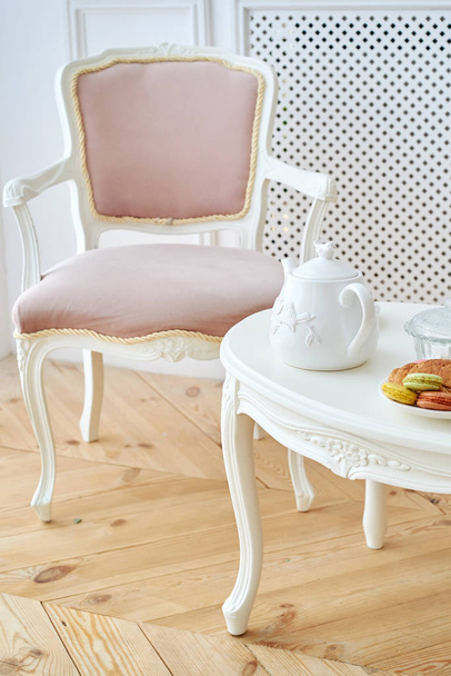 White wooden round table with tea pot, croissant and macaroons in white plate and pink soft chair near. Beautiful modern interior, copy space - Fotoğraf, Görsel