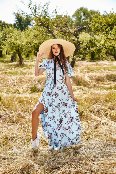 Sexy beautiful young woman fashion model wear long blue stylish dress from summer collection accessory straw hat hot nature garden walk dry grass romance pretty face long brunette hair tan sun shine. - Foto, afbeelding