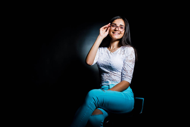 Portrait of an attractive young woman in white top and blue pants sitting posing with her glasses in the dark. - Fotoğraf, Görsel