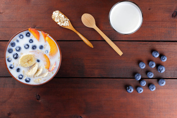 Oatmeal with milk and fruits, blueberries on wooden boards, vegetarian food, useful porridge with banana and peach slices, wooden cutlery, breakfast in vintage style - 写真・画像