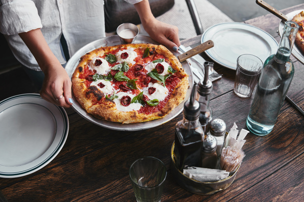 abgeschnittene Aufnahme einer Frau, die einen Teller mit Pizza Margherita bei sich trägt, der im Restaurant auf dem Tisch serviert wird - Foto, Bild