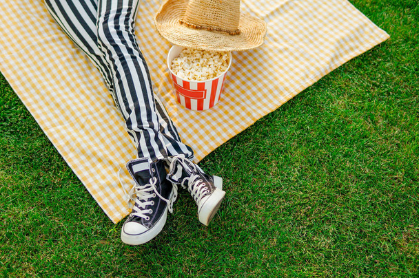Sexy woman legs in striped pants and black snickers eating popcorn on green grass. Food fashion image, top view - Foto, Bild