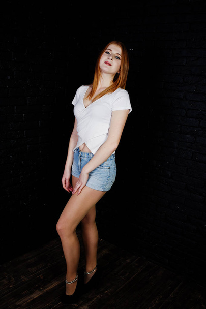 Portrait of a gorgeous redheaded girl in white t-shirt and denim shorts posing in the studio next to the brick wall. - Foto, Imagem