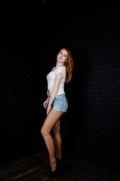 Portrait of a gorgeous redheaded girl in white t-shirt and denim shorts posing in the studio next to the brick wall. - Foto, afbeelding