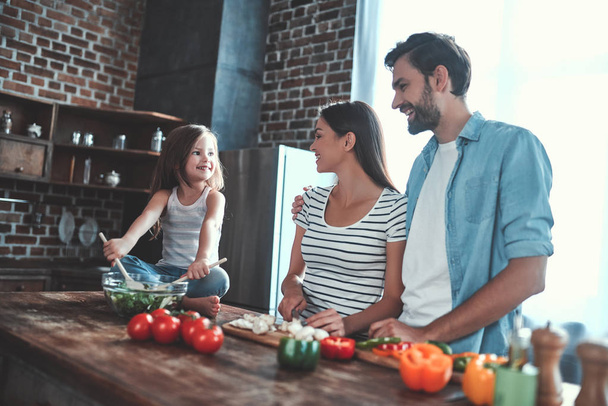 Mom, dad and daughter are cooking on kitchen. Happy family concept. Handsome man, attractive young woman and their cute little daughter are making salad together. Healthy lifestyle. - Photo, Image