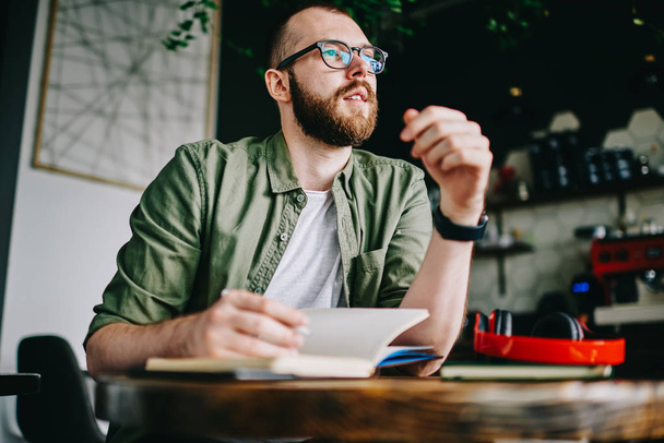 Concentrated student 20 years old in eyeglasses sitting at cafe noting all interesting things of day, young creative hipster guy writing impression review of visiting coffee shop in notebook - 写真・画像