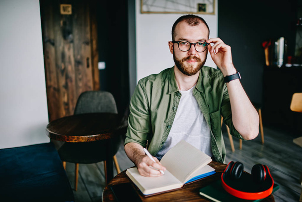 Young hipster male student in eyeglasses writing essay for homework bearded man thinking about tomorrow exam and looking aside, handsome creative author sitting at cafe interior with notebook - Fotografie, Obrázek