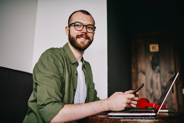 Portrait of handsome hipster man distracted from work on laptop, freelancer holding mobile phone for chatting and using bluetooth connection for share smartphone multimedia files with netbook - Fotó, kép