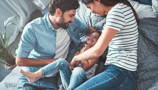 Happy family is having fun in bedroom. Dad, mom and little daughter are spending time together at home while lying in bed and laughing. - 写真・画像
