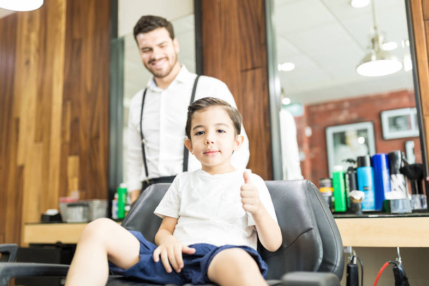 Cool boy showing thumbs up while sitting on chair at salon - Φωτογραφία, εικόνα