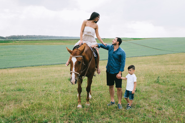 mother riding horse, father and son standing on field - Фото, изображение