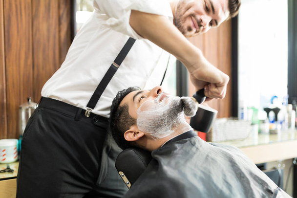 Male barber rubbing shaving foam on client's face with brush in shop - Fotoğraf, Görsel
