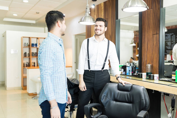 Sonriente joven peluquero ofreciendo asiento al cliente en el salón
 - Foto, Imagen