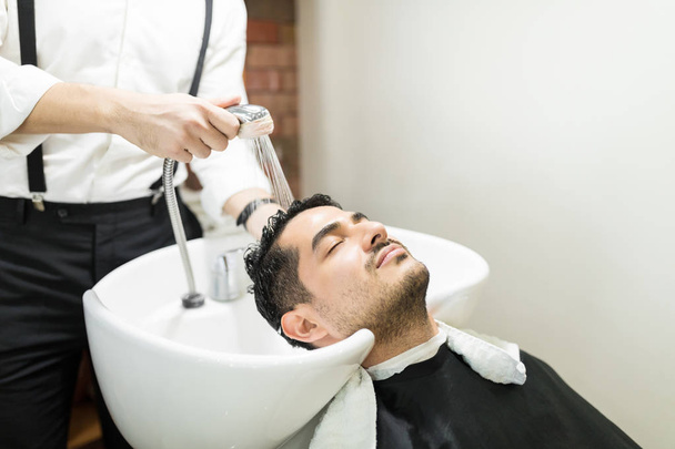 entspannter junger Mann mit geschlossenen Augen lehnt am Waschbecken, während sich der Friseur im Salon die Haare wäscht - Foto, Bild